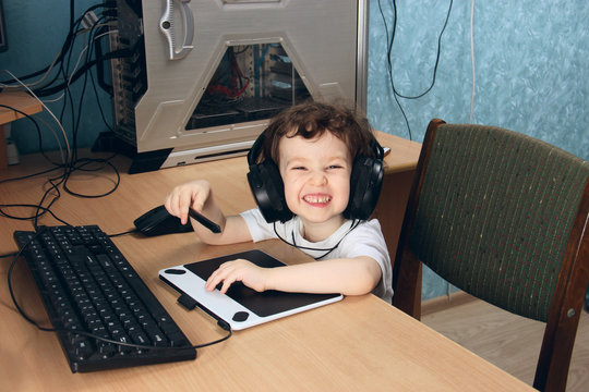 Little 2 3 year old baby girl in white clothers draws at the home computer in graphics drawing tablet. The child is holding a pen and laughs. On the head are huge headphones