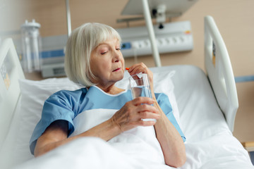 selective focus of senior woman lying in bed, holding glass of water and taking pill in hospital