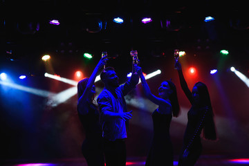  Young friends dancing with glasses of champagne in hands. Against lighting devices as background. Young people's friends are dancing.