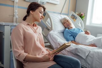 beautiful daughter reading book to sick senior mother lying in bed in hospital
