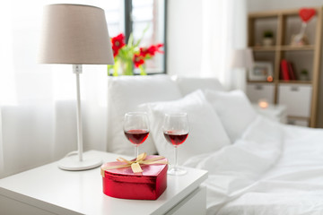 valentines day, love and romantic concept - heart shaped gift box and two glasses of red wine on bedroom nightstand at home
