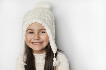 Happy cute child girl in a white winter hat and a white knitted sweater laughs on a white background. Real positive emotions. Smiling beautiful face. 