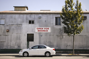 Open for business sign on the side of a corrugated building with a car parked on the street.