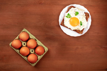 An egg cooked sunny side up on a toast and a carton of eggs, shot from above on a dark rustic wooden background with copy space