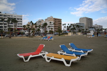 Kolorowe komfortowe łóżka plażowe stoją na środku plaży w Larnace, Cypr, w tle promenada porośnięta palmami, inne leżaki, spacerujący turysta, rząd wysokich hoteli, błękitne niebo