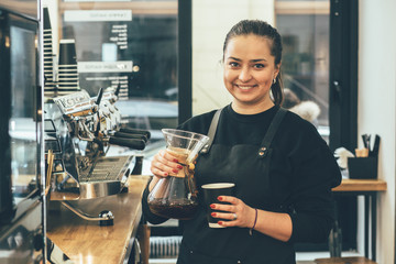 Positive friendly smiling female barista in black uniform preparing coffee drink. Skillful staff...