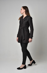 full length portrait of a brunette girl wearing long black coat, standing pose in side profile on grey studio background.