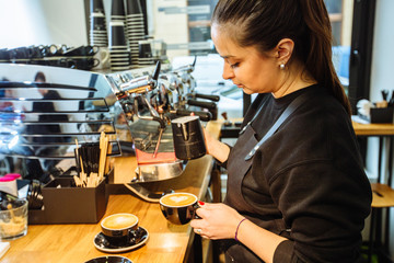 Young brunette caucasian woman barista pouring milk to make cappuccino hot drink. Small local business work in cafe. Skillful staff and real work concept.