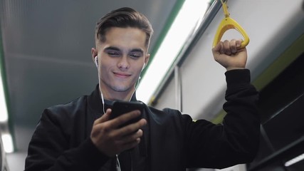 Portrait of attractive men in headphones holds the handrail, watching video on smartphone in public transport. City lights background - Powered by Adobe