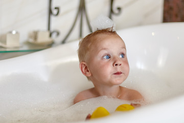 Cute caucasian baby boy with navy blue eyes in the bathtube, with foam bubbles on his head and around. Funny face expression. Indoors, close up portrait of pretty baby, white background, copy space.