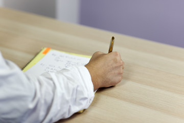 The man' hands on white medical outfit write with roller pen on the paper on wooden table. Indoors, copy space.