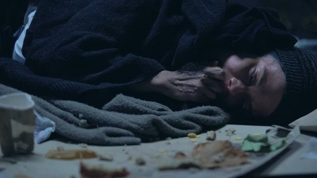 Hungry vagabond lying on ground with garbage and greedily eating orts, shelter
