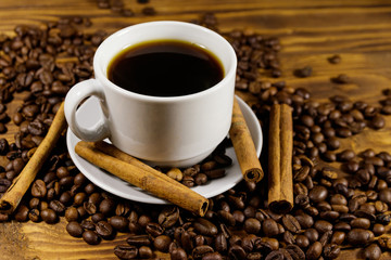 Cup of coffee, roasted coffee beans and cinnamon sticks on wooden table