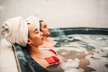 Young asian woman sit in jacuzzi with her european friend. They chill and keep eyes closed. Models wear swimsuits. - obrazy, fototapety, plakaty