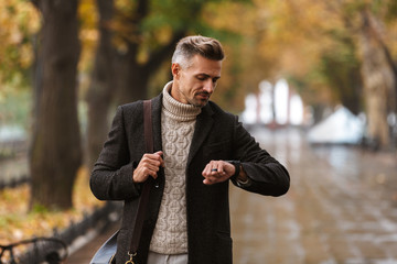 Photo of handsome man 30s wearing warm clothes walking outdoor through autumn park, and looking at wrist watch