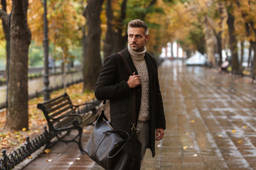 Photo of adult stylish man 30s wearing warm clothes walking outdoor through autumn park, and looking aside