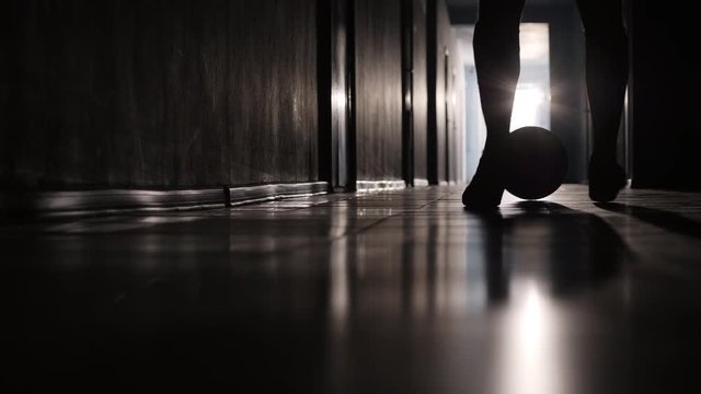 Low section shot of silhouette of legs of man in shorts running along hallway and dribbling soccer ball 