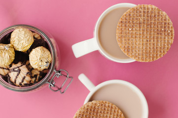 Two coffee cups with dutch caramel waffles and jar with sweets and cookies, top view, pink background