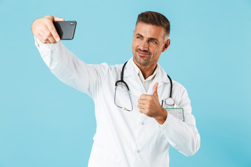 Photo of young medical doctor wearing white coat and stethoscope taking selfie on smartphone, standing isolated over blue background