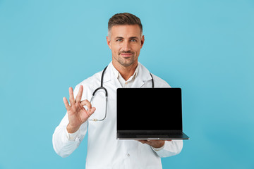 Photo of optimistic man wearing white medical coat and stethoscope holding laptop, standing isolated over blue background