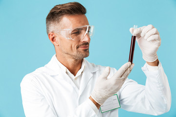 Portrait of medical doctor wearing white uniform holding test tubes with blood, standing isolated over blue background