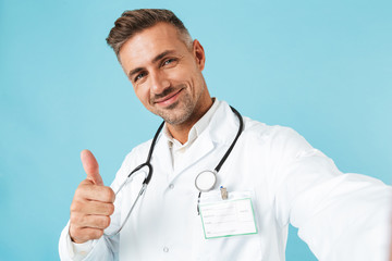 Portrait of happy medical doctor with stethoscope taking selfie photo, while standing isolated over blue background