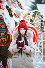 Young beautiful cheerful brunette girl walking in the winter at the fair. Positive winter portrait on the background of Christmas decorations on the city street.