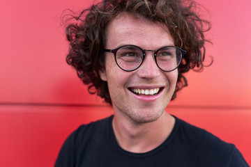 Closeup portrait of handsome smiling male posing for advertisement on red wall outdoor wearing black t-shirt and round eyeglasses with copy space for your informational text. People emotions