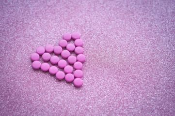 Obraz na płótnie Canvas pink pills laid out in shape of a heart on glitter pink background. coloured drugs. concept - heart disease, heart disorders and drugs, cardiology, valentines, love,