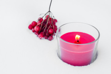 a red candle stands in the snow. On the background of the candle is a branch of the guelder rose