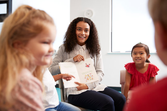 Over shoulder view of female teacher showing a picture in a book to a group of kindergarten children sitting on chairs in a classroom, close up
