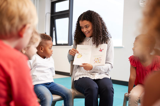 Young female teacher showing a picture in a book to kindergarten children sitting on chairs in a circle in the classroom, close up, selective focus