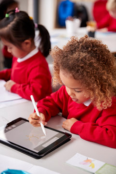 Elevated View Of Young Schoolgirl Wearing School Uniform Sitting At Desk In An Infant School Classroom Using A Tablet Computer And Stylus, Close Up, Vertical