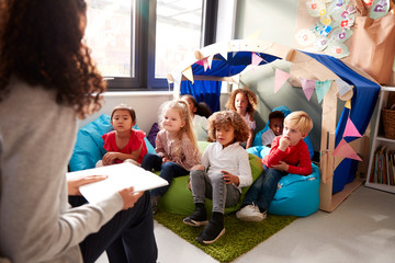 Female infant school teacher sitting on a chair reading a book to a group of children sitting on...