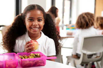 Young black schoolgirl sitting at a table smiling and holding an apple in a kindergarten classroom during her lunch break, close up