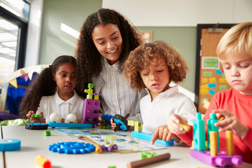 Female teacher sitting at table in play room with three kindergartne children constructing, selective focus - Powered by Adobe