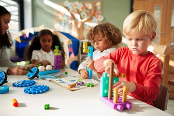 Naklejka premium Infant school boy sitting at a table using educational construction toys with his classmates, close up