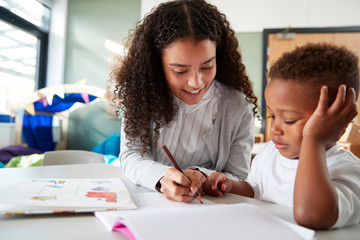 Female infant school teacher working one on one with a young schoolboy, sitting at a table writing...