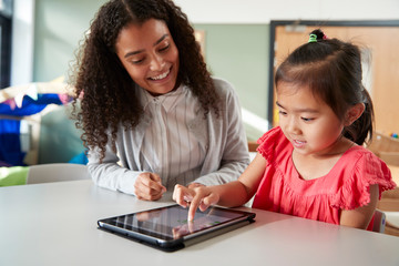 Female infant school teacher working one on one with a Chinese schoolgirl, sitting at a table in a...