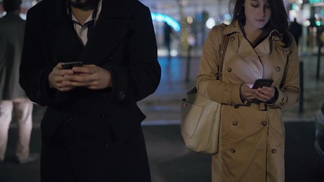 Couple using smartphones and crossing road at night. Cropped view of young man and woman in trench coats using cell phones and walking on urban city street. Technology concept