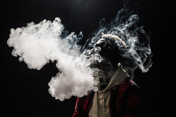 Feeling relaxed. Portrait of afro guy holding vape device and surrounded by cloud of smoke isolated on black background - Powered by Adobe