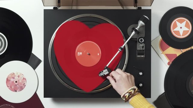A red heart vinyl record turntable on a white table with plates. Included gramophone, torque. The hand of a girl DJ with bright accessory puts a stylus with a needle on a vinyl record. Valentine's Day