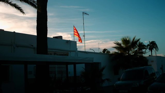 Catalan flag, Senyera, waving on a pole on the roof of a building against a blue sky near sunset hour with palm trees nearby