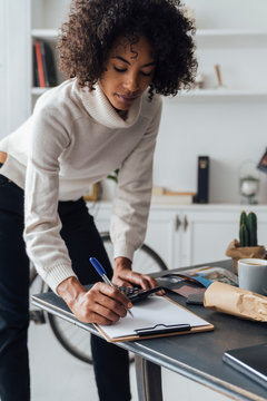 Freelancer standing at her desk, using calculator, taking notes