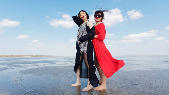 Happy smiling pretty Chinese girls having fun on sunny beach. Stylishly dressed in red dress and black dress, sunglasses, best young friends hand in hand and hug in love. 