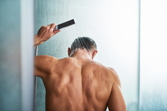 Young man using handheld shower head while washing himself in bathroom