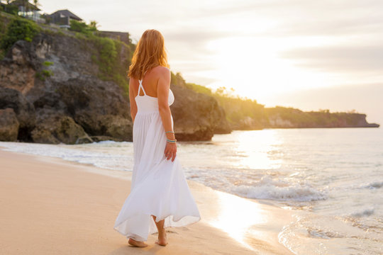 Relaxed woman in white dress walking on the beach in evening