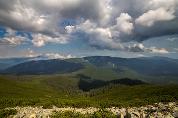 Bright white puffy clouds lit by sun spreading against blue summer sky over green mountain ridge.