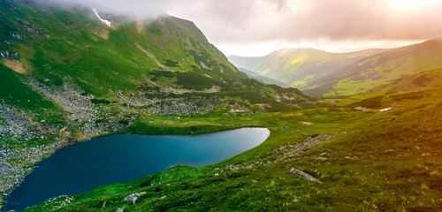 Fototapeta premium Fantastic wide peaceful view of blue lake in green valley and small tourist tents at rocky mountain with patches of snow covered with low clouds. Beauty of nature, tourism and traveling concept.