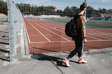 Athletic guy in headband dressed in black sport clothes with backpack on his shoulders walks along the playground fence on a sunny day